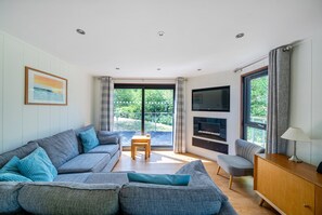 A modern living room with a grey sofa, a wooden coffee table, a TV mounted on the wall, and a view of greenery outside. - Beckington Lodge -  A Peaceful, rural retreat on the outskirts of Frome, near Longleat Safari Park a (Beckington)