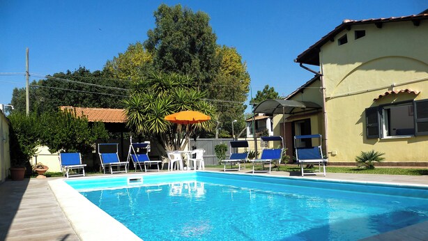 Piscina com espreguiçadeiras e guarda-sol, rodeada por vegetação e edifícios.