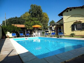 A swimming pool with lounge chairs and an umbrella, surrounded by greenery and buildings.