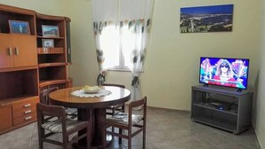 Uma sala de jantar com uma mesa redonda, cadeiras, um armário de madeira com prateleiras, uma televisão sobre um suporte e uma janela com cortinas.