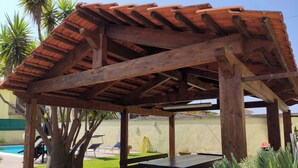 Estrutura de pérgola de madeira com telhado de azulejos, mesa e cadeiras por baixo.
