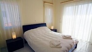 Una cama bien tendida en una habitación de hotel, con una mesita de noche negra, una lámpara, cortinas traslúcidas y suelo de baldosas.