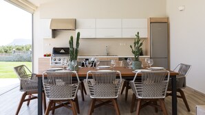 A modern kitchen with a dining area, featuring a wooden table and wicker chairs, with a view of greenery outside. - Aelia Residence, Wellness & Leisure with 100m² Heated Pool, Whirlpool, Sauna&Gym (Rethymnon)
