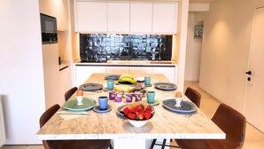 A modern kitchen with a dining table set for four, featuring a marble countertop, a window with a view of trees, and a kitchen island with a sink. - Maramar Westend Luxury Apartment with Sea View (Middelkerke)