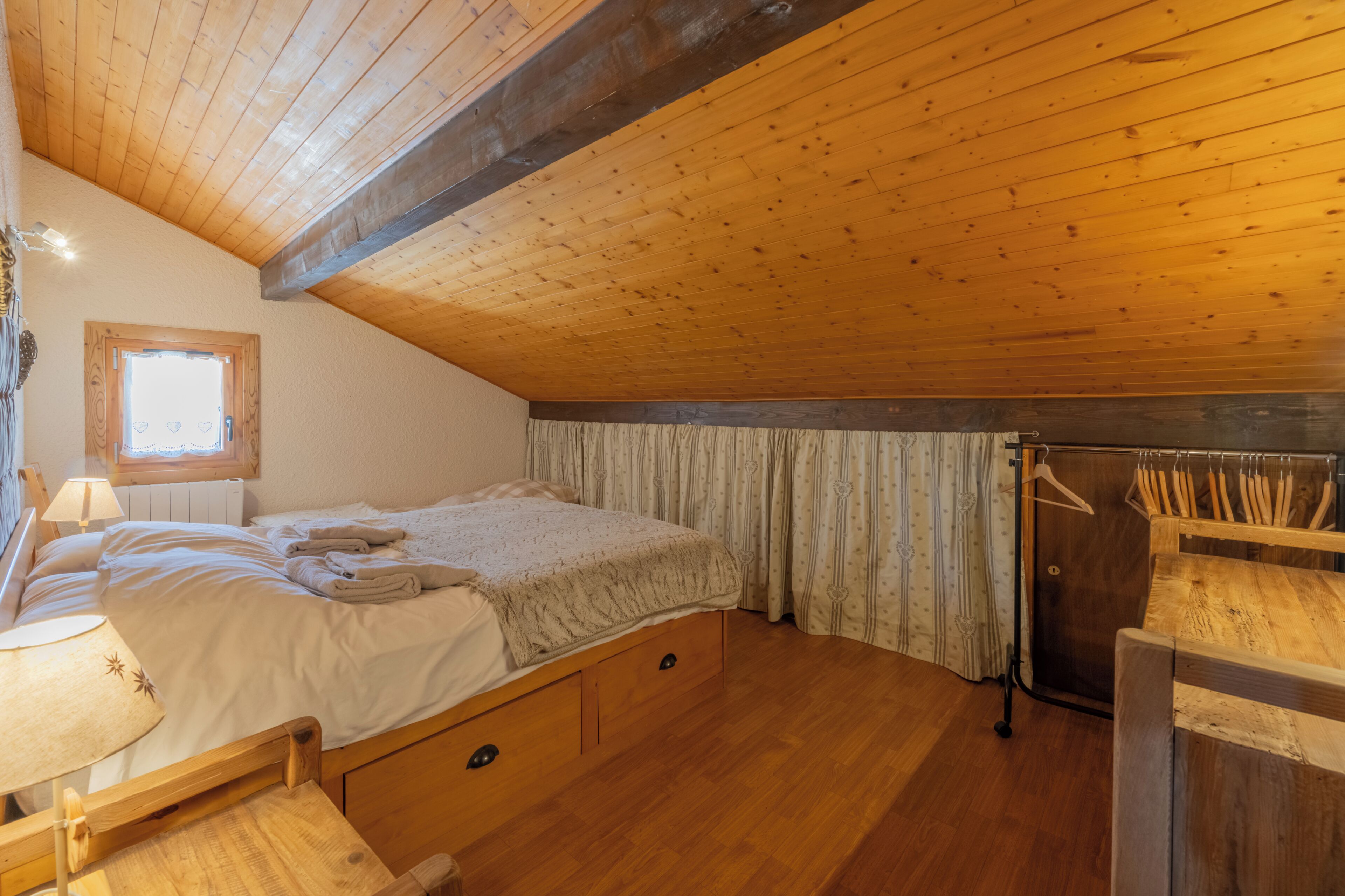 A bedroom with a wooden ceiling, a bed, a window, a radiator, and a wooden chair.