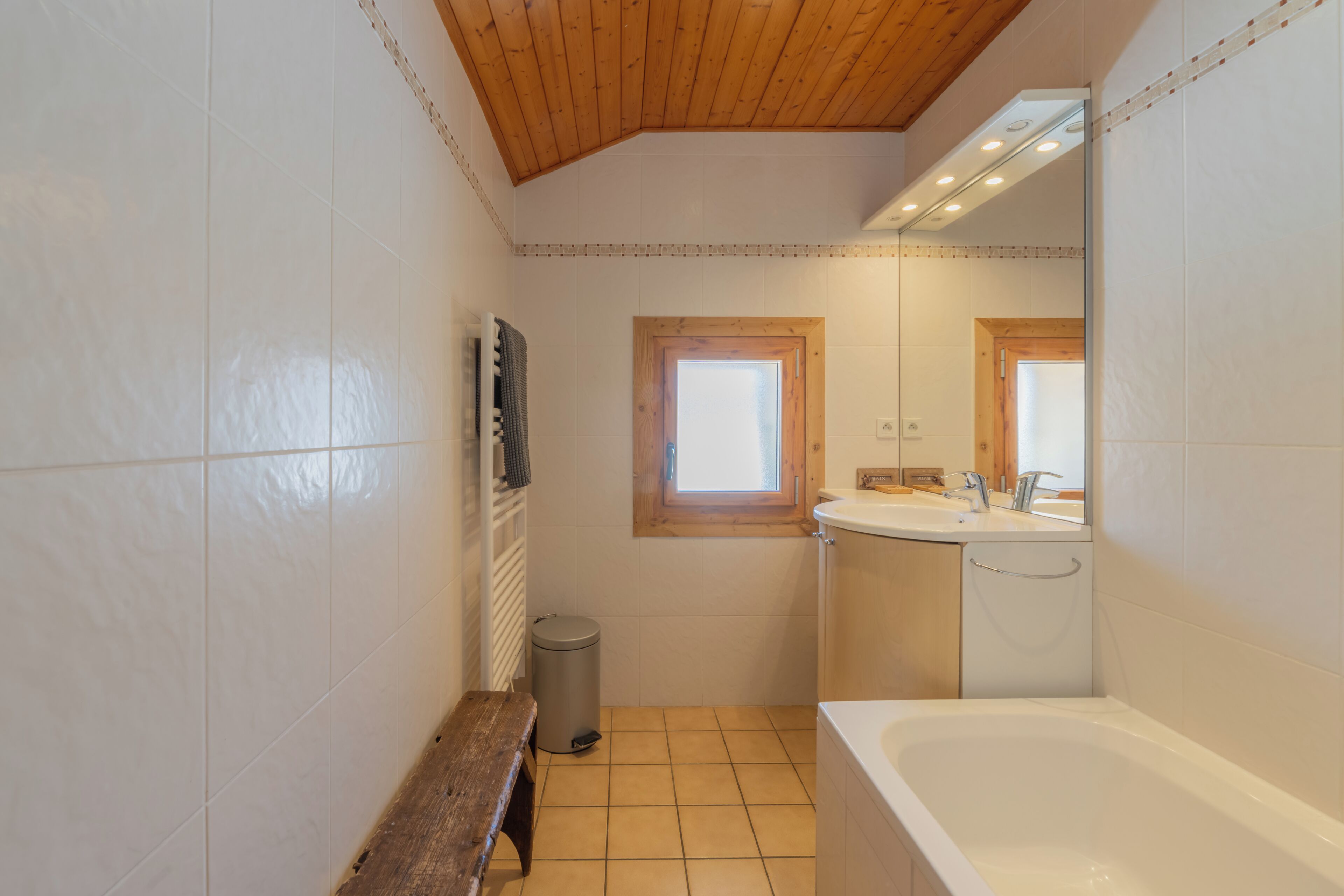 Une salle de bain avec un plafond en bois, des carreaux blancs, un banc, des toilettes, un lavabo et une baignoire.