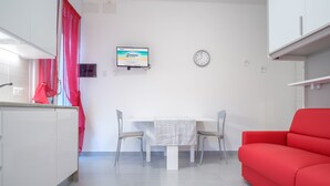 Une cuisine moderne avec des meubles blancs, un rideau rouge, un téléviseur fixé au mur et une table à manger avec deux chaises.