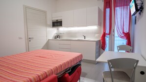 Une pièce de taille modeste comprenant un lit, un bureau, une chaise, un coin cuisine et une fenêtre avec des rideaux.