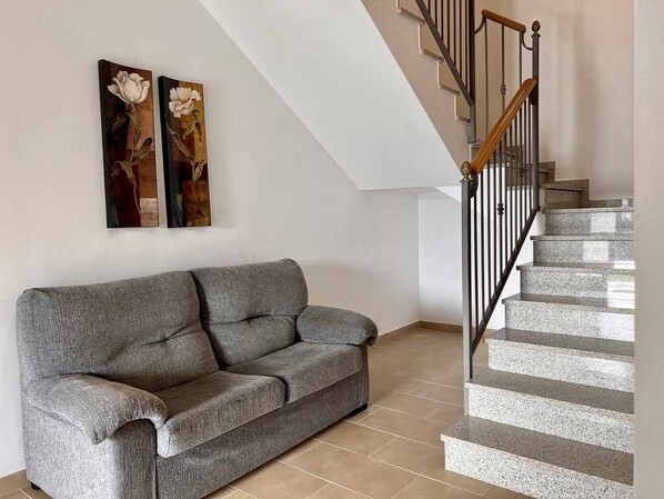 Een woonkamer met een grijze bank, een trap met een houten leuning, en twee aan de muur bevestigde schilderijen.