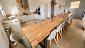 A modern kitchen with a long wooden table and white chairs, a built-in oven, and a large window. - Mas Ambros House with infinity pool and barbecues in Calonge, Girona Costa Brava (Calonge)