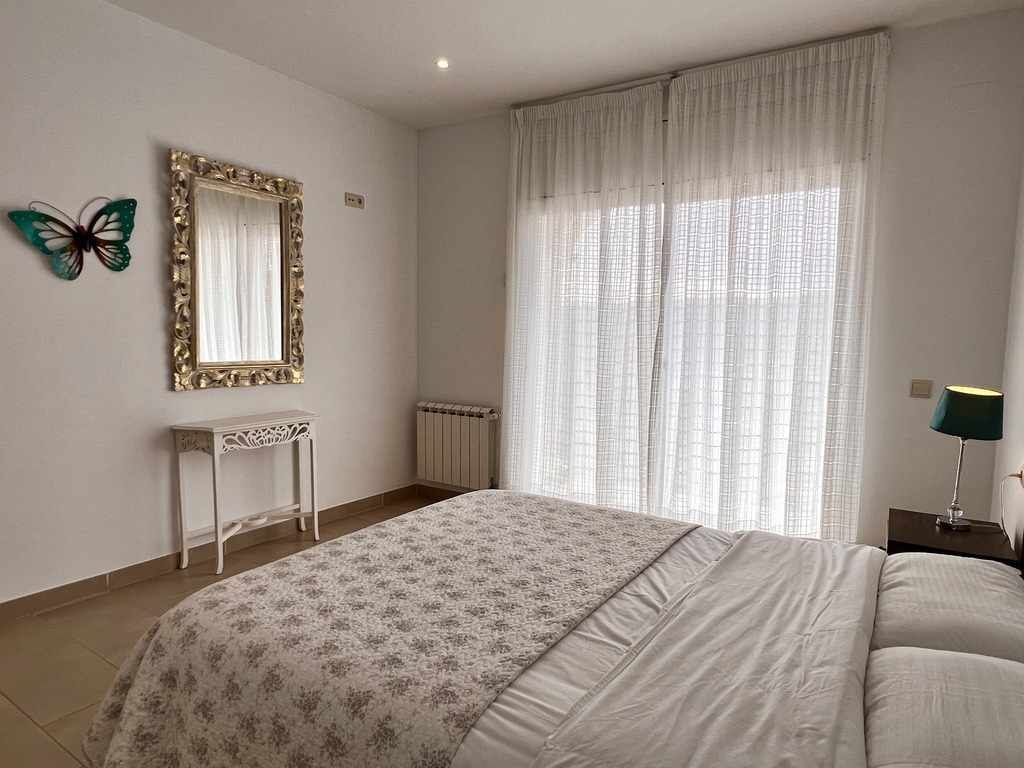 Une chambre à coucher comprenant un lit, une table de chevet, une lampe, une fenêtre avec des rideaux, un radiateur et un miroir fixé au mur.