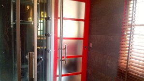 Ein Badezimmer mit Duschkabine aus Glas, einer Tür mit rotem Rahmen und einem Fenster mit Jalousien.