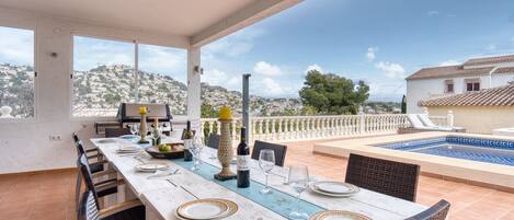 A dining area with a long table set for a meal, overlooking a swimming pool and a scenic hillside view.