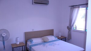 Una cama bien tendida con una colcha azul y blanca, una mesita de noche con lámpara, un aire acondicionado de pared y una ventana con cortinas.
