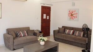 Una sala de estar con dos sofás, una mesa de centro con un jarrón de flores, un equipo de aire acondicionado, una puerta y un ventilador de pared.