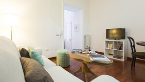 Ein Wohnzimmer mit Sofa, Hocker, Couchtisch, Bücherregal, Fernseher und einer Tür zu einem weiteren Raum.