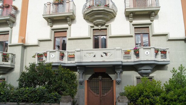 Ein Gebäude mit einem zentralen Eingang, Balkonen mit Blumenkästen und einer Stein-Fassade.