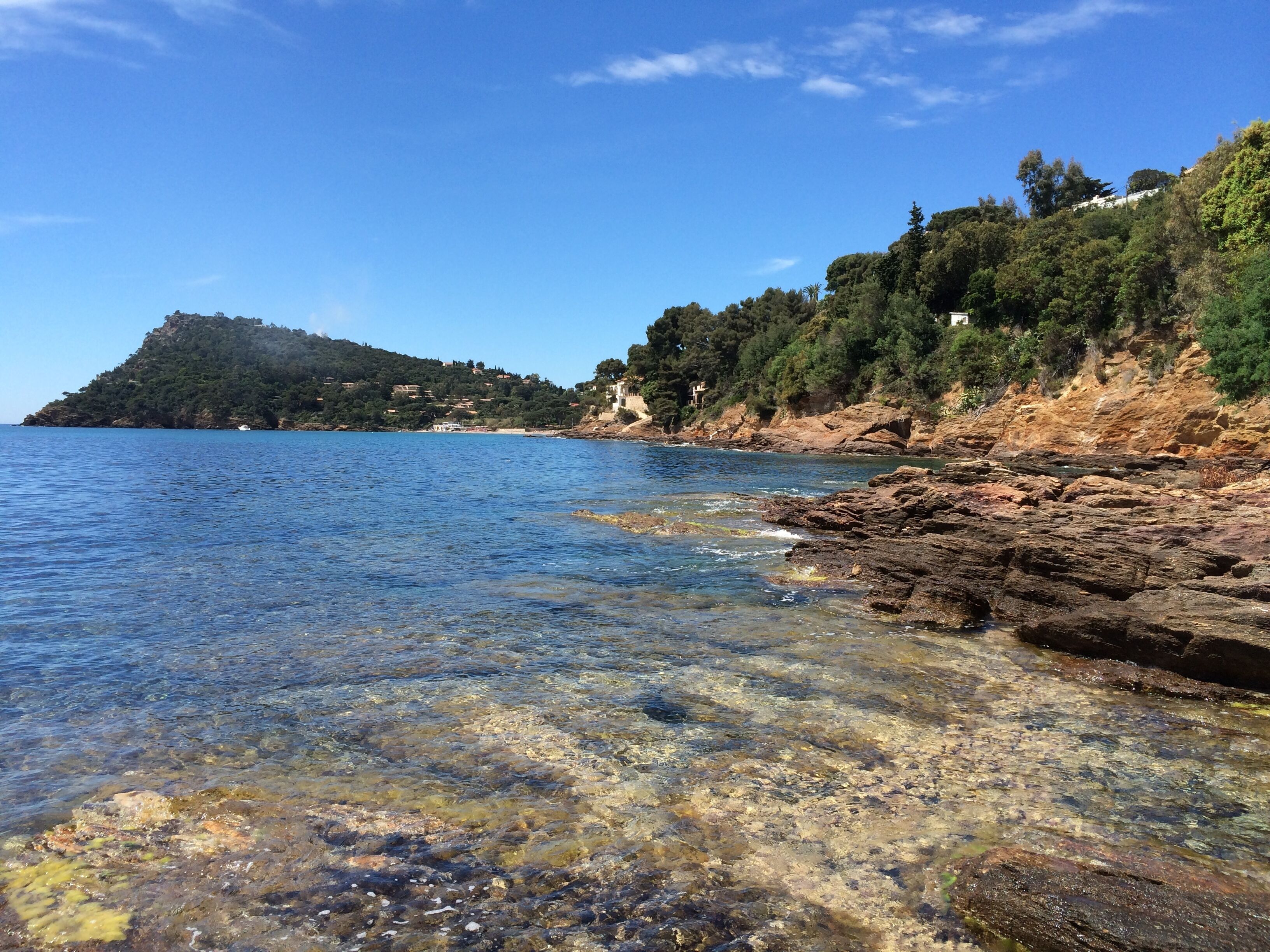 A coastal landscape with clear water, rocky shore, and a hill with buildings.
