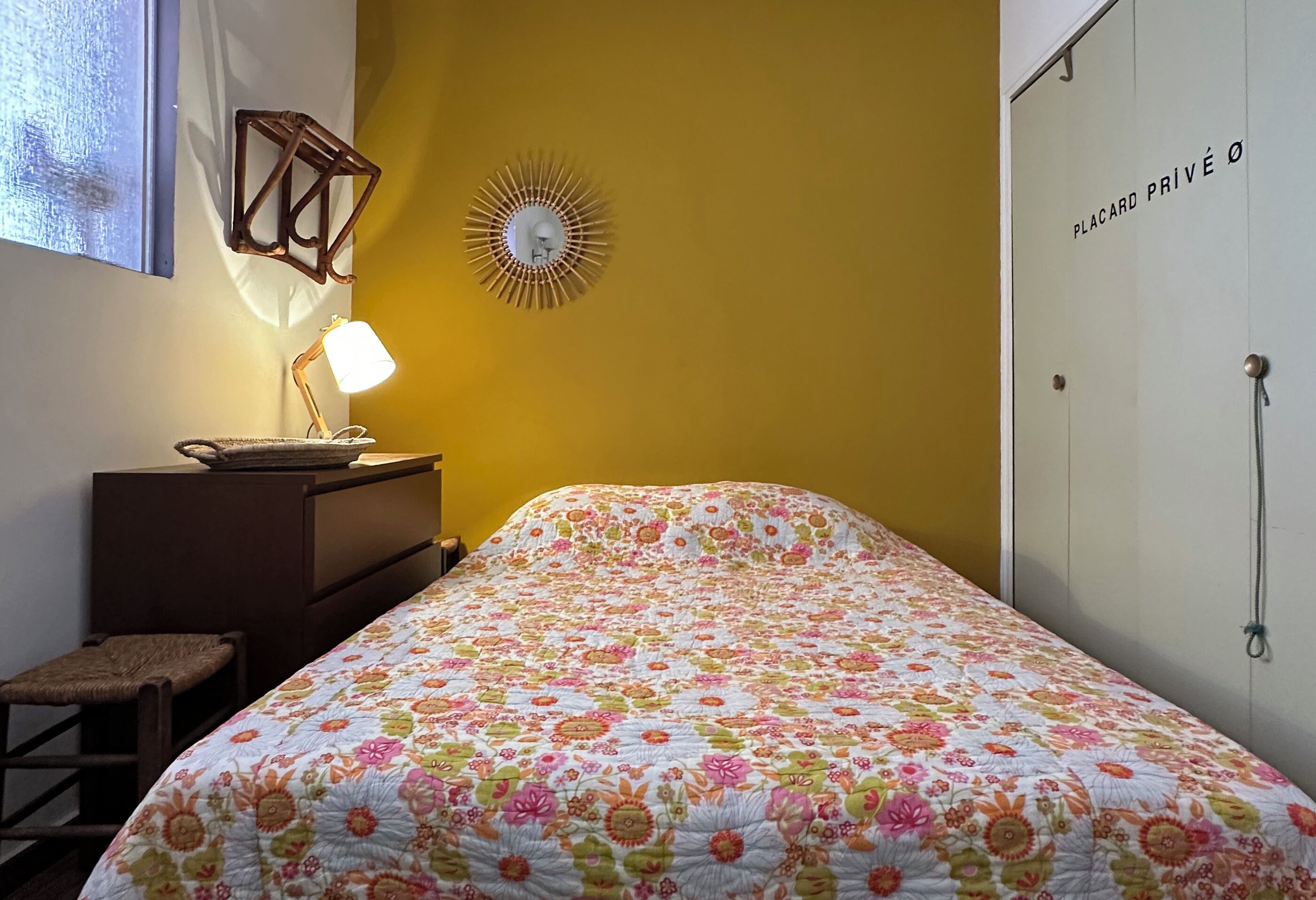 Un dormitorio con colcha estampada, lámpara de noche, un reloj de pared y un armario con una puerta que dice 'PLACARD PRIVÉ Ø'.