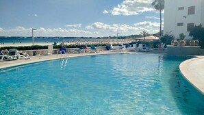 En swimmingpool med liggestole og parasoller, en strand i baggrunden, og en bygning med hvide facader.