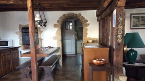A rustic kitchen with stone walls, wooden beams, a stone archway, a wooden bench, a table with a bowl, and a green lamp.