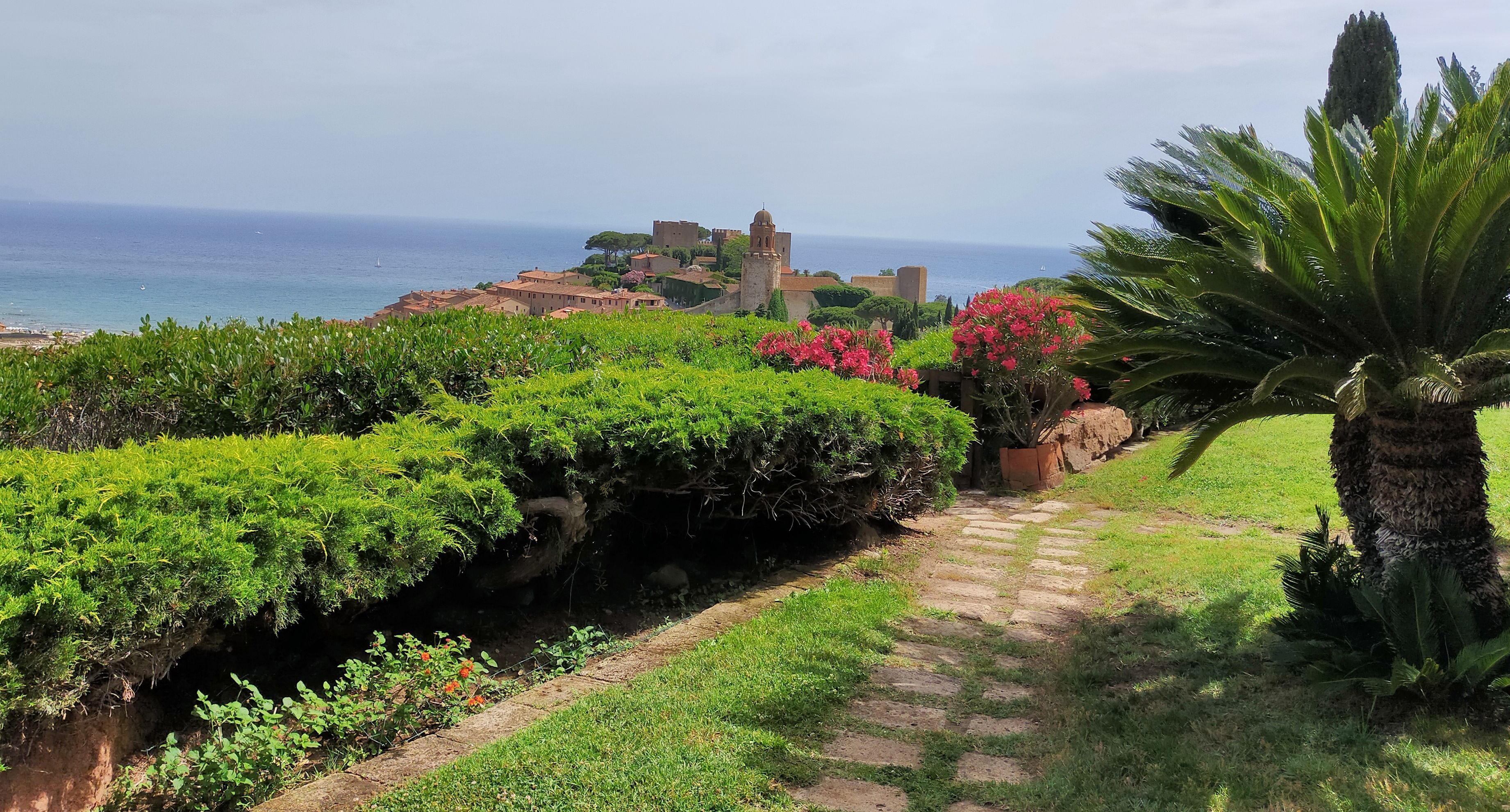 A pathway leading to a coastal town with historical buildings, lush greenery, and a view of the sea.