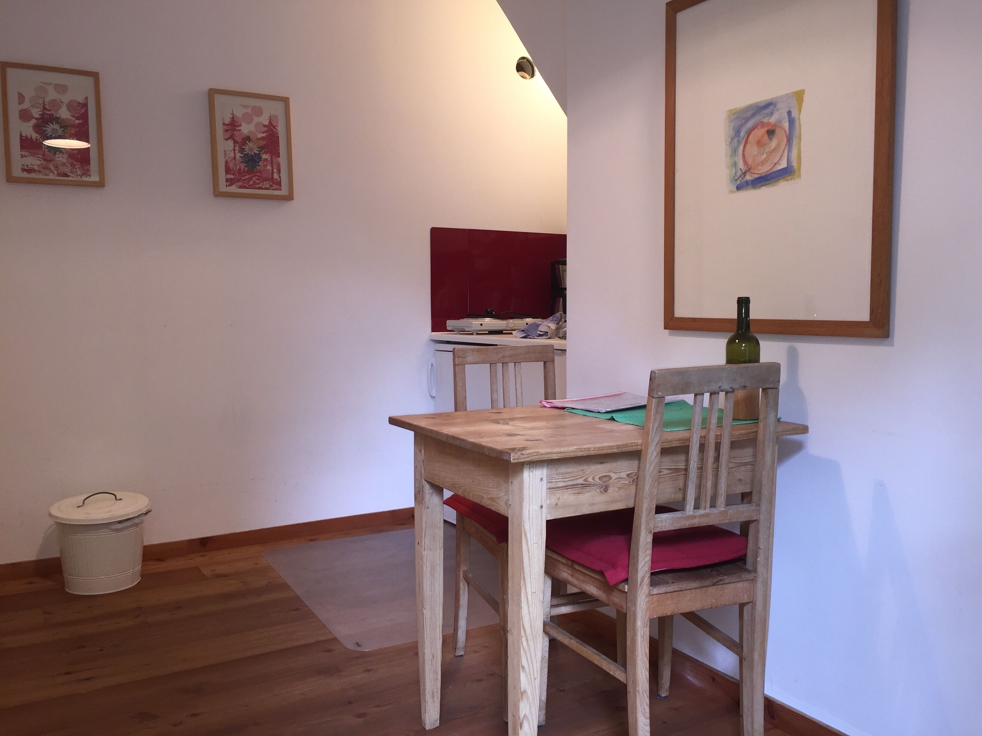 A wooden dining table with a red cushion, a wooden chair, a white trash bin, and a framed picture on the wall.