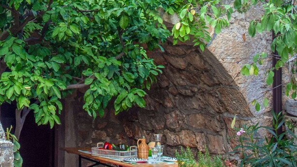 Un espace de restauration en plein air, avec un mur en pierre en arrière-plan, une table en bois dressée avec des assiettes, des verres et un pichet, et un banc agrémenté d’un coussin rouge.