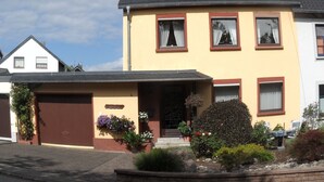A two-story house with a garage, a front porch with potted plants, and a small garden area. - Quiet Non smoking apartment (approx 80sqm.) Located right on the edge. (Holzerath)