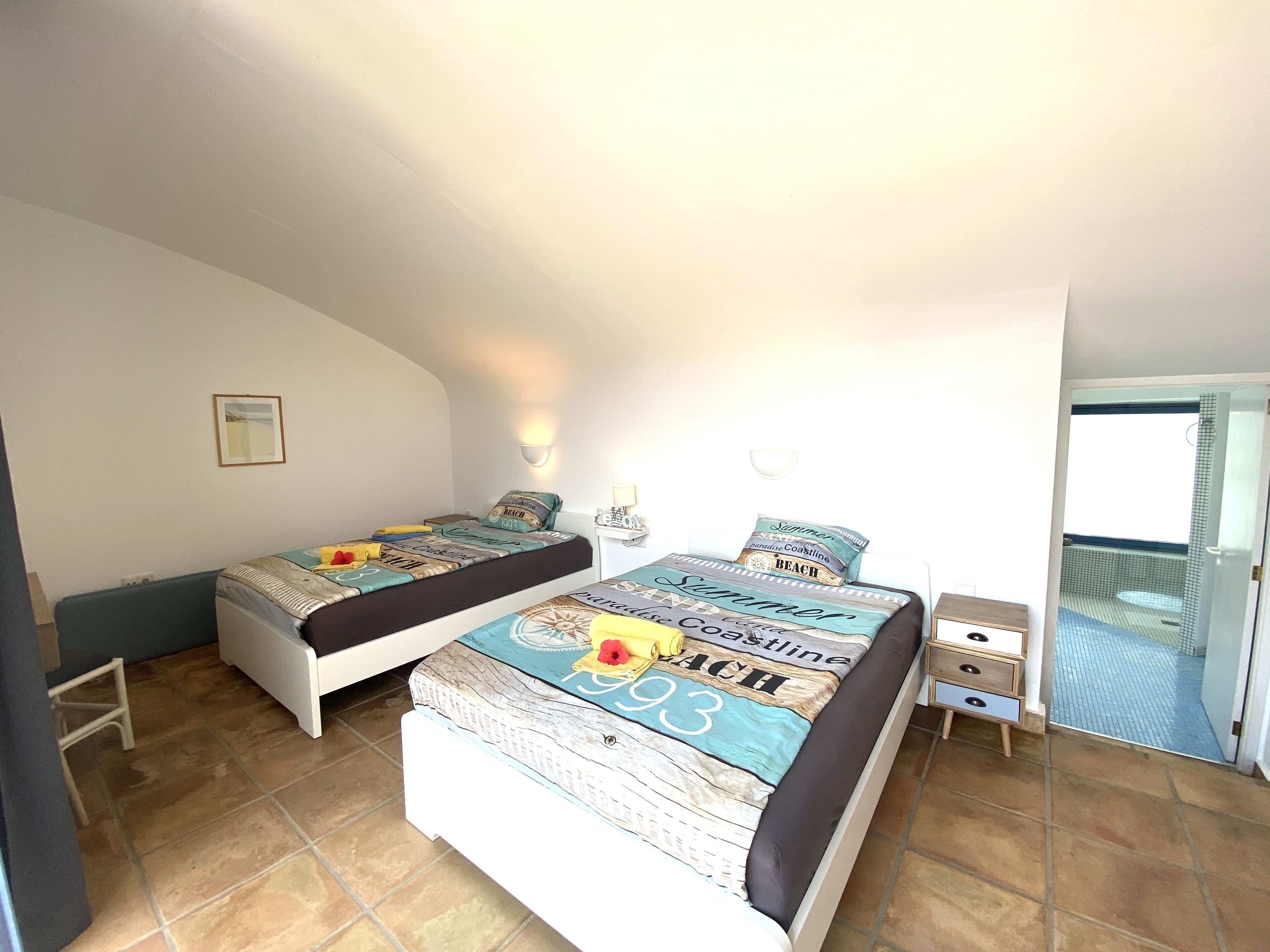 Un dormitorio con dos camas individuales, una mesita de noche pequeña con una lámpara, un cuadro en la pared y vistas a un balcón a través de una puerta abierta.