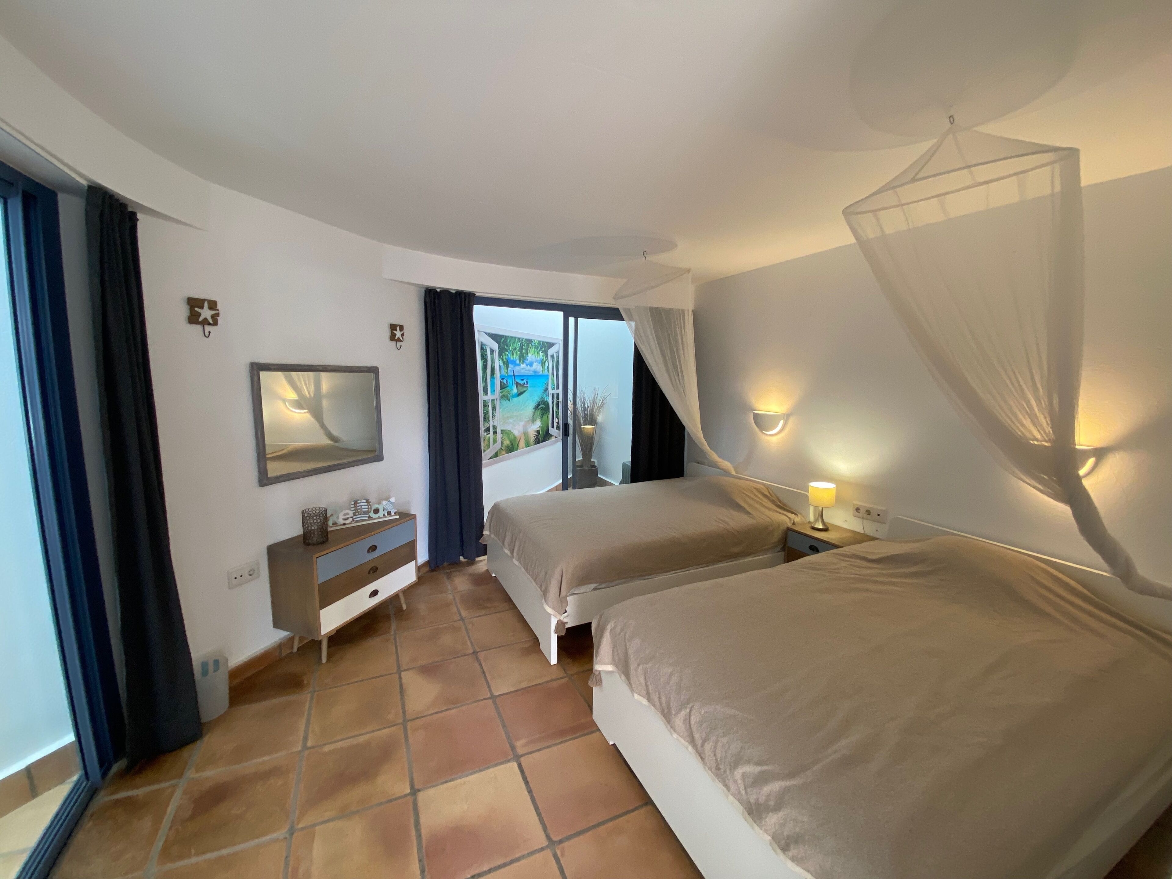 A hotel room with two beds, a nightstand with a lamp, a dresser, a mirror, and a view of a beach through an open door.
