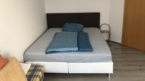 Una cama individual con cabecera oscura, ropa de cama gris y una manta azul.