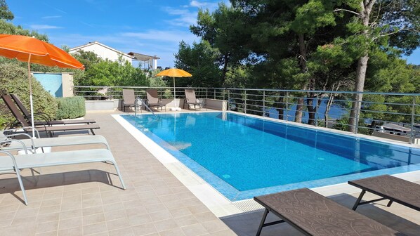 A swimming pool with lounge chairs and umbrellas, surrounded by trees and a building.