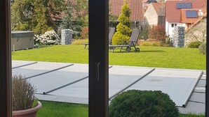 A view through a glass door of a residential area with solar panels, a lawn, and a church steeple in the background. - Comfortable apartment with pool for 2-6 people (110 sqm) (Ranschbach)