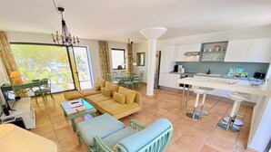 A modern living room with a sofa, a dining area with chairs, a kitchen with white cabinets, and a large window with curtains. - Casa Barbara, private pool, internet, close to the ocean, sheltered courtyard/patio (Costa Calma)