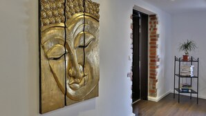 A hallway with a large gold-colored artwork on the wall, a brick wall, a black door, and a metal shelf with books and a potted plant.