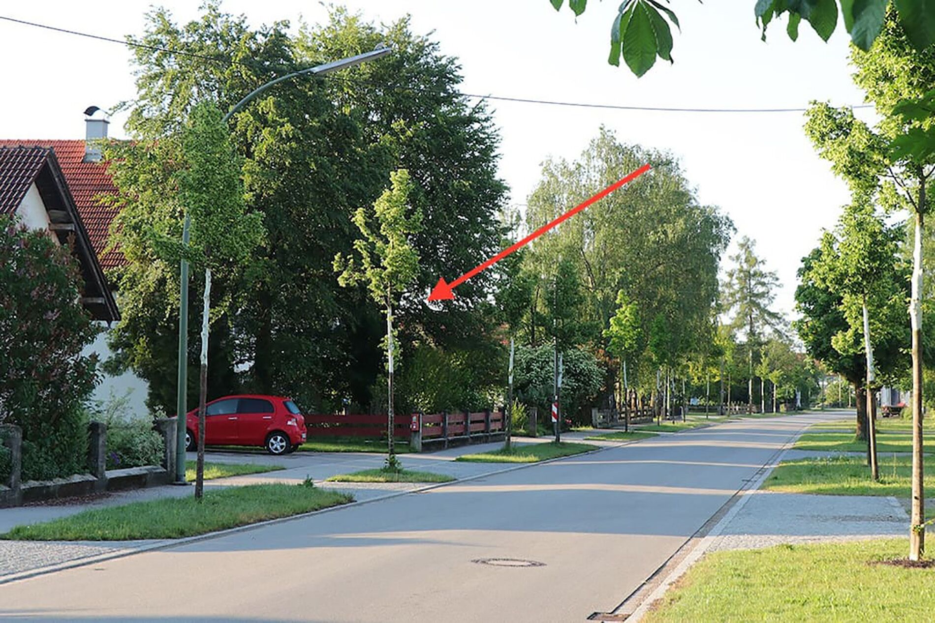 Une rue paisible dans une banlieue résidentielle, avec une voiture rouge garée sur le bord, bordée d’arbres et flanquée d’un bâtiment en briques rouges.
