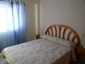 Quarto com cabeceira de madeira, colcha florida, mesa de cabeceira com abajur e janela com cortinas azuis.