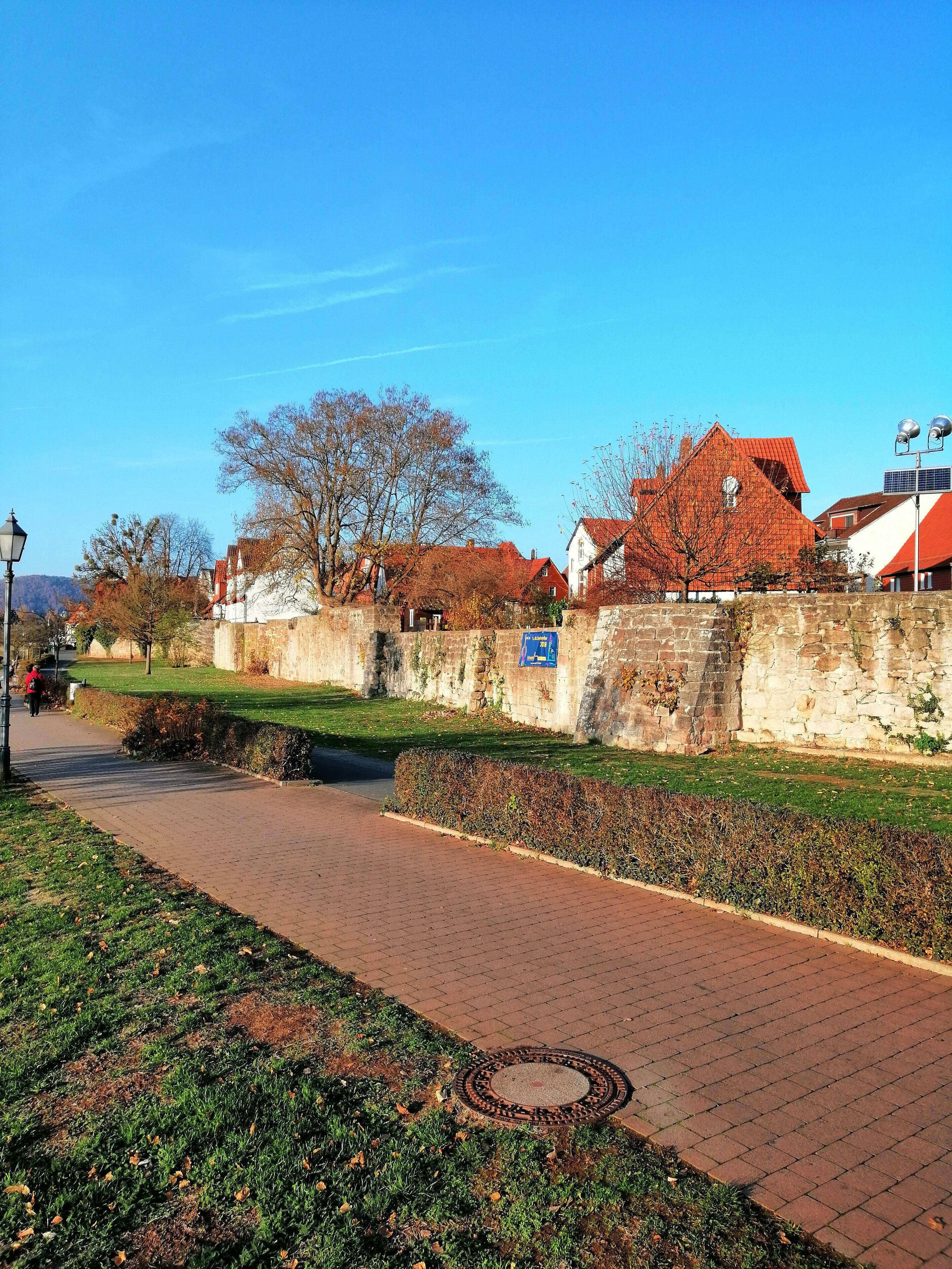 A paved pathway alongside a stone wall with a small bridge, leading to traditional buildings with red roofs.