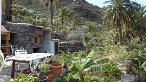 Un edificio de piedra rústico con un patio que incluye una mesa y sillas, rodeado de plantas en macetas y palmeras.