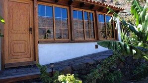 Un edificio con puerta de madera y ventanas amplias, rodeado de plantas y un muro de piedra.