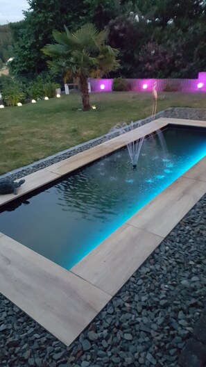 Une piscine rectangulaire avec une fontaine au centre, entourée d’une bordure de galets et d’une terrasse en bois.