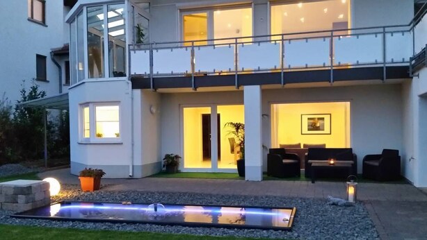 Een modern huis met twee verdiepingen, een balkon, een goed verlichte entree en een klein terras met een waterpartij.