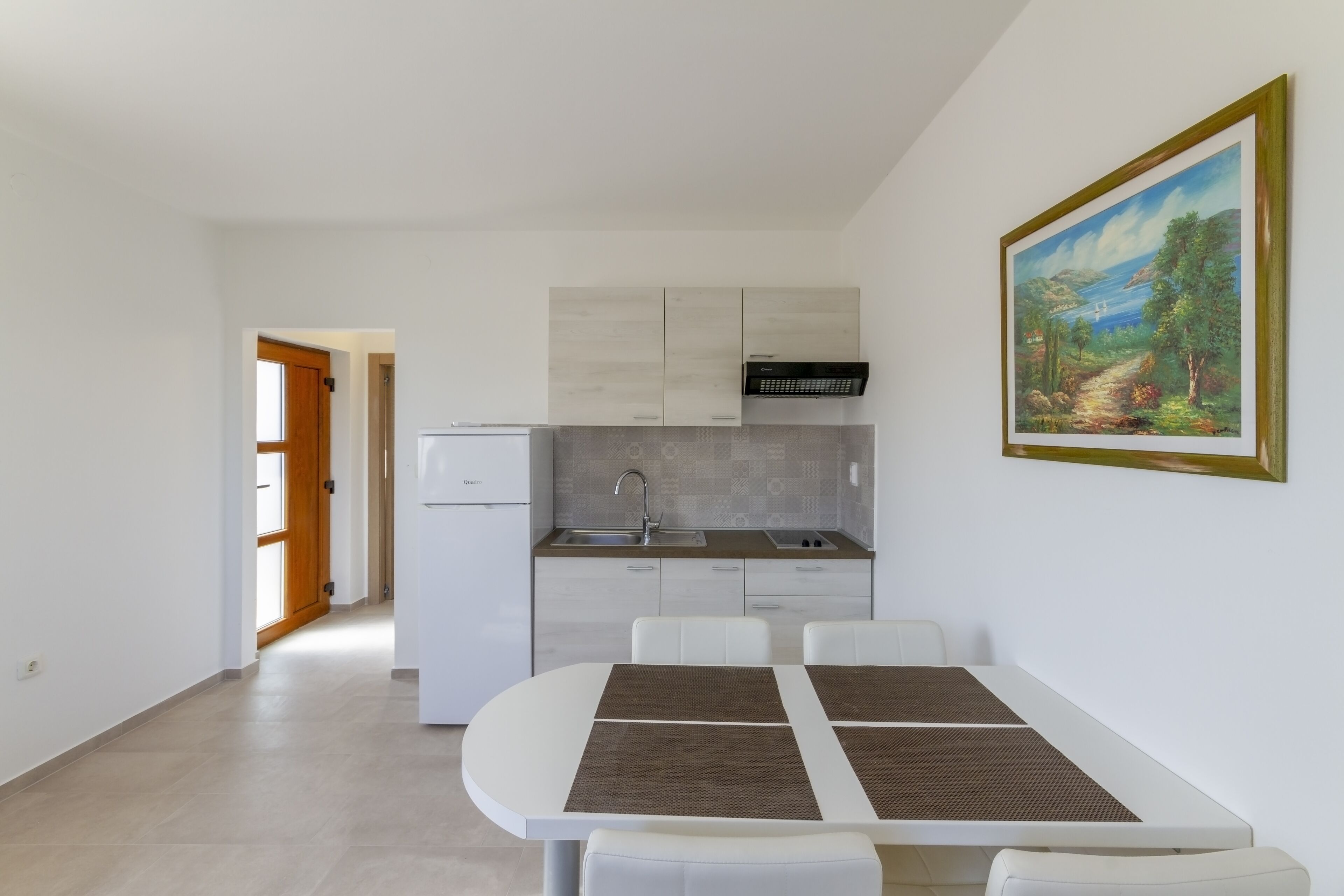 A modern kitchen with white cabinets, a built-in oven, and a sink. There is a dining area with a white table and chairs, and a painting on the wall.
