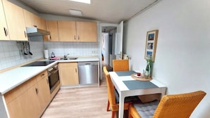A compact kitchen with wooden cabinets, a dishwasher, and a dining area with wicker chairs and a table. - Daub's island oasis/apartment with terrace also possible with dog (Borkum)