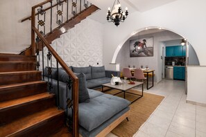 A modern living room with a grey sofa, wooden staircase, and a kitchen area with teal cabinets.