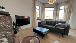 Una sala moderna con televisor de pantalla plana, un sofá gris, una mesa de centro de madera y un ventanal con cortinas.