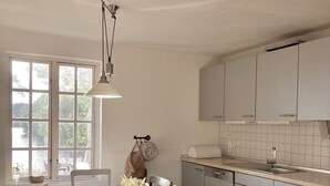 A kitchen with a long wooden table, white chairs, a hanging light fixture, and a window with a view of greenery. - Vacation home "Backhaus" directly on the lake at Gut Wittmoldt (Wittmoldt)
