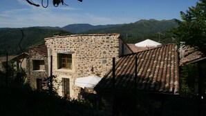 A stone building with a tiled roof, surrounded by greenery and mountains in the background.