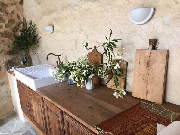 A wooden counter with a white sink, two vases with white flowers, a wooden cutting board, and a wall-mounted light fixture.
